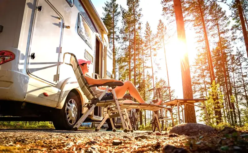A camper relaxing beside an RV among tall pines at sunrise